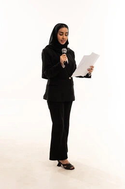 إعداد وتحضير أفكار الخطاب بشكل مسبق، قراءة الملاحظات والمعلومات، بدء التدريب الصوتي، بورتريه لامرأة عربية سعودية خليجية مبتسمة ترتدي البدلة الرسمية السوداء تقف باستقامة و تمسك بيدها المايكروفون و الأوراق, خلفية بيضاء