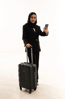 التجهيز لرحلة عمل, التأكد من موعد الطائرة عبر الهاتف, الاستعداد لبدء العطلة خارج البلاد, بورتريه لامرأة عربية سعودية خليجية محجبة ترتدي البدلة الرسمية السوداء تقف باستقامة و تقوم بجر حقيبة الامتعة, خلفية بيضاء