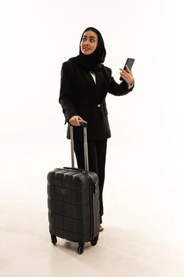الاستعداد للسفر في رحلة عمل,  دمج التقنية والتكنولوجيا في الحياة العملية، بورتريه لامرأة عربية سعودية خليجية محجبة ترتدي البدلة الرسمية السوداء تقف باستقامة و تقوم بجر حقيبة الامتعة و تحمل بيدها الموبايل, ايماءات السعادة والسرور، خلفية بيضاء