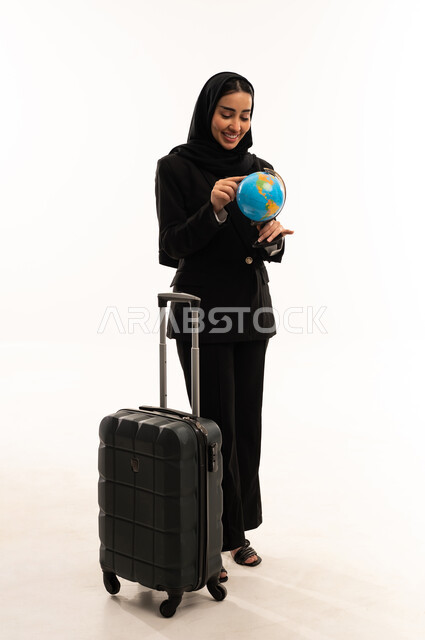 Determining the itinerary, research and exploration around the world, the concept of tourism and travel, taking leisure tours, a portrait of a veiled Saudi Arabian woman from the Gulf wearing a black formal suit, standing upright, pulling a suitcase and holding a globe, white background