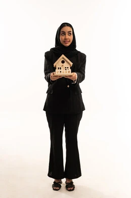 The pursuit of owning a new home, buying and selling real estate, a portrait of a veiled Saudi Arabian woman from the Gulf region wearing a black formal suit, standing upright and holding a small wooden model of a house with gestures of joy and hope, white background