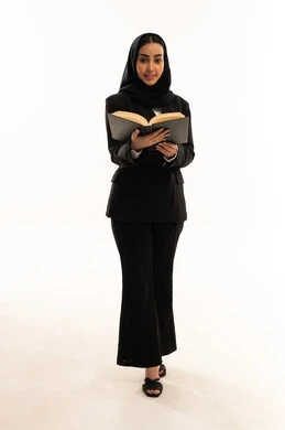 شغف التعرف على الكتب المحتلفة, حب المطالعة والاستكشاف, قضاء أوقات الفراغ بالتثقيف والتعليم, بورتريه لامرأة عربية سعودية خليجية مبتسمة محجبة ترتدي البدلة الرسمية السوداء تقف باستقامة و تحمل كتاب, ثقافة التعلم الذاتي, خلفية بيضاء