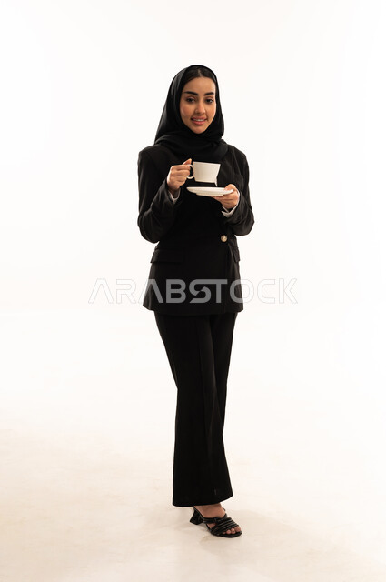 الحيوية والنشاط عند شرب الكافيين، لحظات الهدوء والراحة, الاستمتاع والاحساس بالدفء, بورتيه لامرأة عربية سعودية خليجية محجبة ترتدي البدلة الرسمية السوداء تقف باستقامة و تحمل بيدها فنجان من الشاي، خلفية بيضاء