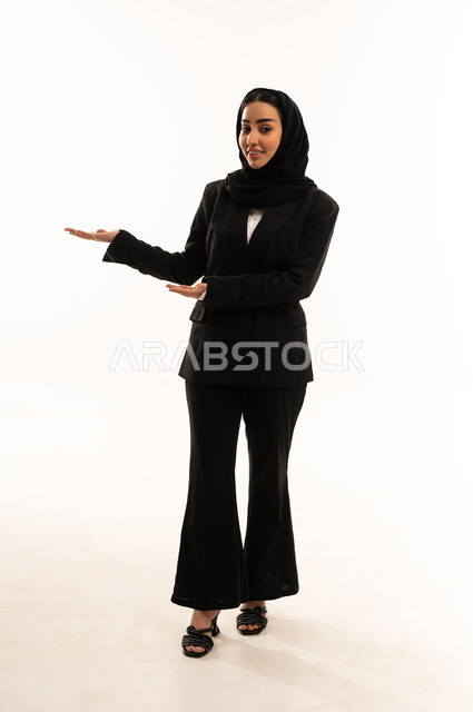 Indicating something through gestures of presence, emphatic gestures, and identifying the object opposite; a portrait of a veiled Saudi Arabian woman from the Gulf region wearing a black formal suit, standing upright and gesturing with her hand in a warm and welcoming manner, against a white background.