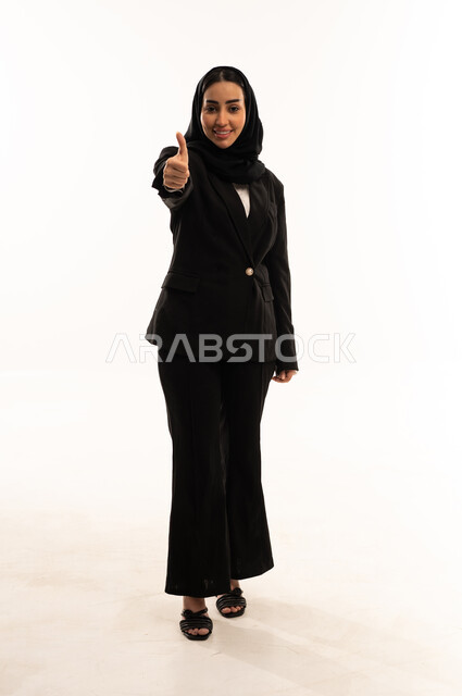 Admiration, satisfaction, and success; facial and hand gestures indicating approval and acceptance; a portrait of a veiled Saudi Arabian woman from the Gulf region wearing a black formal suit, standing upright and raising her thumb in a thumbs-up gesture; white background.