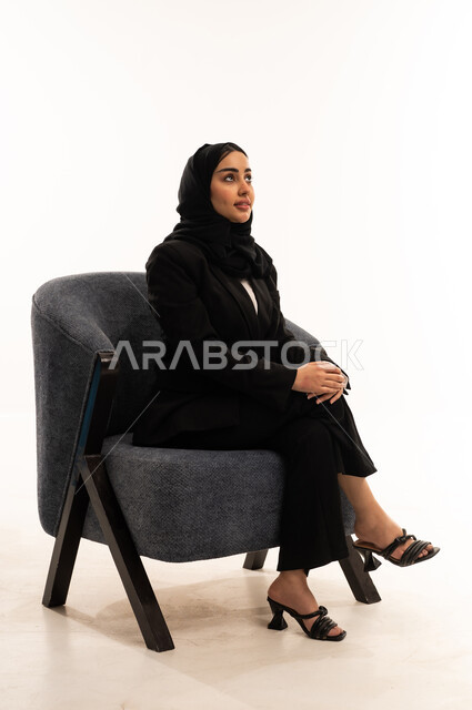 مفهوم الجلوس والراحة، الاستمتاع بالأجواء الهادئة، مفهوم الأناقة والاهتمام بالمظهر الخارجي، التأمل والتخطيط للمستقبل، بورتيه لامرأة عربية سعودية خليجية محجبة ترتدي البدلة الرسمية السوداء تجلس على كرسي مريح, خلفية بيضاء