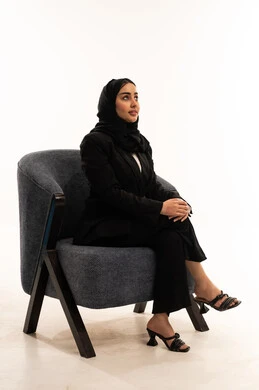 The concept of sitting and relaxing, enjoying a tranquil atmosphere, the concept of elegance and attention to outward appearance, contemplation and planning for the future: a portrait of a veiled Saudi Arabian woman from the Gulf region wearing a black formal suit, sitting in a comfortable chair, against a white background.