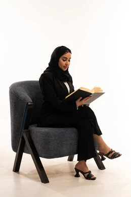 تنمية مهارات القراءة, الاستمتاع بمطالعة الكتب والقصص والروايات, قضاء الوقت بالتعلم والتثقف، بورتيه لامرأة عربية سعودية خليجية محجبة ترتدي البدلة الرسمية السوداء تجلس على كرسي مريح و تحمل بيدها كتاب، ثقافة التعلم الذاتي, خلفية بيضاء