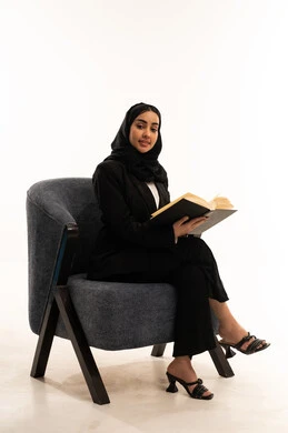 المطالعة والثقافة والإستطلاع،  قضاء الوقت بالقراءة والتعلم والتثقف، بورتيه لامرأة عربية سعودية خليجية محجبة ترتدي البدلة الرسمية السوداء تجلس على كرسي مريح و تحمل بيدها كتاب، ثقافة التعلم الذاتي, خلفية بيضاء