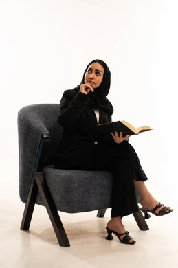 التعلم والتثقيف من خلال قراءه الكتب الهادفه، المطالعة والثقافة والإستطلاع، بورتيه لامرأة عربية سعودية خليجية محجبة ترتدي البدلة الرسمية السوداء تجلس على كرسي مريح و تحمل بيدها كتاب، ثقافة التعلم الذاتي, خلفية بيضاء