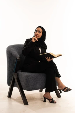 كتب وروايات هادفه ومسليه, المطالعة والثقافة والإستطلاع، قضاء الوقت بالقراءة والتعلم والتثقف، بورتيه لامرأة عربية سعودية خليجية محجبة ترتدي البدلة الرسمية السوداء تجلس على كرسي مريح و تحمل بيدها كتاب، ثقافة التعلم الذاتي, خلفية بيضاء