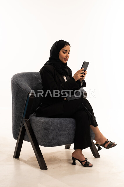Browsing social media, spending quality time on your mobile with friends, connecting and chatting with family and friends via mobile apps; a portrait of a smiling, veiled Saudi Arabian woman wearing a black formal suit, sitting in a comfortable chair and holding a mobile phone; white background.