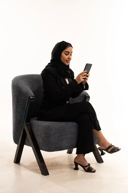 تصفح مواقع التواصل الاجتماعي، قضاء وقت ممتع عبر الجوال مع الأصحاب، التواصل والدردشة مع الأهل والأصدقاء عبر الهاتف, بورتيه لامرأة عربية خليجية سعودية محجبة مبتسمة ترتدي البدلة الرسمية السوداء تجلس على كرسي مريح و تحمل بيدها موبايل, خلفية بيضاء