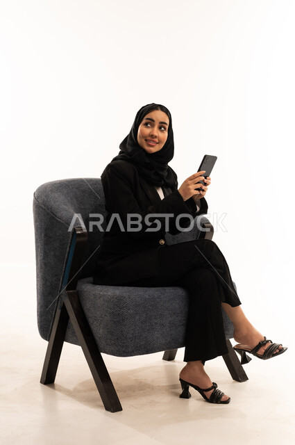 قضاء وقت ممتع عبر الجوال مع الأصحاب، تصفح مواقع التواصل الاجتماعي، التواصل والدردشة مع الأهل والأصدقاء عبر الهاتف, بورتيه لامرأة عربية خليجية سعودية محجبة مبتسمة ترتدي البدلة الرسمية السوداء تجلس على كرسي مريح و تحمل بيدها موبايل, خلفية بيضاء