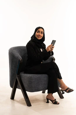 قضاء وقت ممتع عبر الجوال مع الأصحاب، تصفح مواقع التواصل الاجتماعي، التواصل والدردشة مع الأهل والأصدقاء عبر الهاتف, بورتيه لامرأة عربية خليجية سعودية محجبة مبتسمة ترتدي البدلة الرسمية السوداء تجلس على كرسي مريح و تحمل بيدها موبايل, خلفية بيضاء