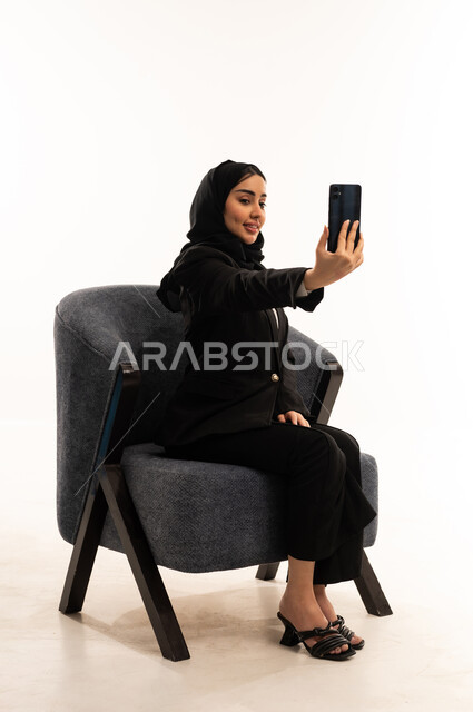 توثيق اللحظات و الذكريات, إجراء مكالمات الفيديو عبر الانترنت، بورتيه لامرأة عربية سعودية خليجية محجبة ترتدي البدلة الرسمية السوداء تجلس على كرسي مريح و تحمل بيدها موبايل و تلتقط صورة, خلفية بيضاء