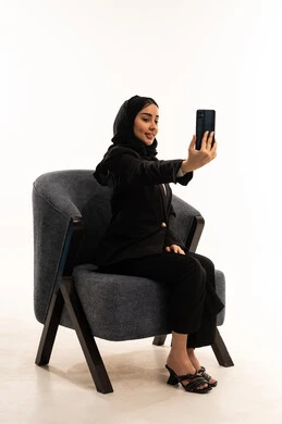 توثيق اللحظات و الذكريات, إجراء مكالمات الفيديو عبر الانترنت، بورتيه لامرأة عربية سعودية خليجية محجبة ترتدي البدلة الرسمية السوداء تجلس على كرسي مريح و تحمل بيدها موبايل و تلتقط صورة, خلفية بيضاء