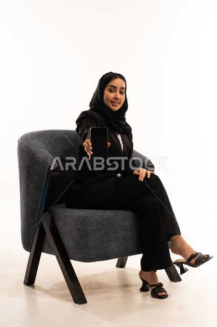 قضاء وقت الفراغ بالتواصل مع الصديقات، عرض مواقع وخدمات الشركة عبر الجوال, جهاز تقني حديث ومتطور, بورتيه لامرأة عربية سعودية خليجية محجبة ترتدي البدلة الرسمية السوداء تجلس على كرسي مريح و تعرض شاشة الموبايل, خلفية بيضاء