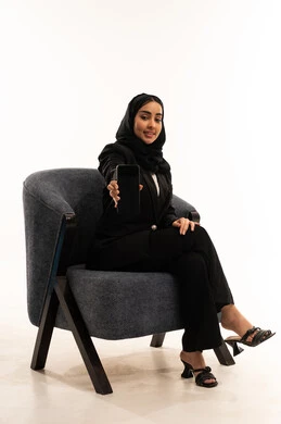 Spending free time connecting with friends, viewing company websites and services via mobile, a modern and advanced technological device, a portrait of a veiled Saudi Arabian Gulf woman wearing a black formal suit, sitting in a comfortable chair and displaying her mobile screen, white background