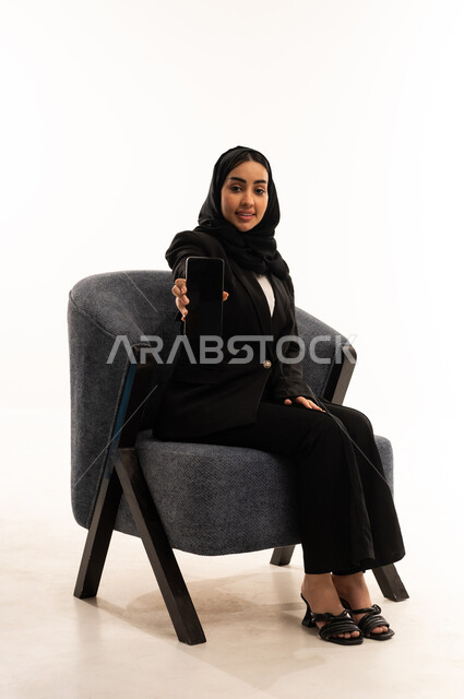 Displaying the company's websites and services via mobile phone, a modern and advanced technological device, a portrait of veiled a Saudi Arabian Gulf woman wearing a black formal suit, sitting in a comfortable chair and displaying her mobile phone screen, white background.
