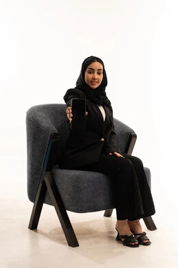 Displaying the company's websites and services via mobile phone, a modern and advanced technological device, a portrait of veiled a Saudi Arabian Gulf woman wearing a black formal suit, sitting in a comfortable chair and displaying her mobile phone screen, white background.