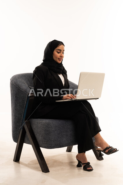 اللعب على اللابتوب, إجراء مكالمات فيديو عبر الإنترنت,  بورتريه لامرأة عربية سعودية خليجية محجبة ترتدي البدلة الرسمية السوداء تجلس على كرسي مريح و تقوم باستخدام جهاز اللابتوب, الاندماج والتركيز لإنجاز المهام عن بعد، خلفية بيضاء