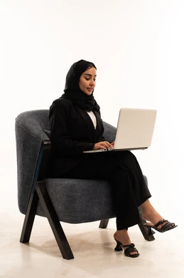 اللعب على اللابتوب, إجراء مكالمات فيديو عبر الإنترنت,  بورتريه لامرأة عربية سعودية خليجية محجبة ترتدي البدلة الرسمية السوداء تجلس على كرسي مريح و تقوم باستخدام جهاز اللابتوب, الاندماج والتركيز لإنجاز المهام عن بعد، خلفية بيضاء