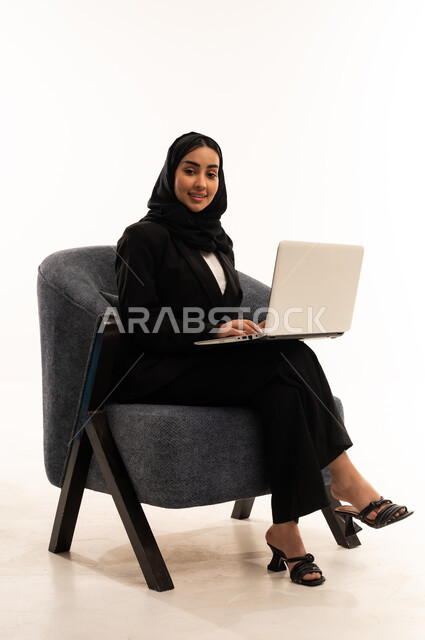 دردشة وتواصل عبر الانترنت, استخدام الاجهزة والتقنيات الحديثة والمتطورة, بورتريه لامرأة عربية سعودية خليجية محجبة ترتدي البدلة الرسمية السوداء تجلس على كرسي مريح و تقوم باستخدام جهاز اللابتوب, الاندماج والتركيز لإنجاز المهام عن بعد، خلفية بيضاء