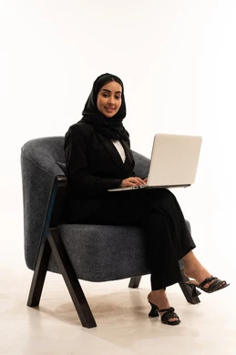دردشة وتواصل عبر الانترنت, استخدام الاجهزة والتقنيات الحديثة والمتطورة, بورتريه لامرأة عربية سعودية خليجية محجبة ترتدي البدلة الرسمية السوداء تجلس على كرسي مريح و تقوم باستخدام جهاز اللابتوب, الاندماج والتركيز لإنجاز المهام عن بعد، خلفية بيضاء