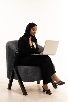 Integration and focus for remote task completion, remote work meeting, online chat and communication, portrait of a veiled Saudi Arabian Gulf woman wearing a black formal suit, sitting in a comfortable chair, using a laptop, and raising her hand in a welcoming gesture, white background