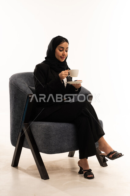 Enjoying the tranquil atmosphere, sipping your favorite beverage, sitting and drinking hot beverages and warming winter herbs, a portrait of a veiled Saudi Arabian woman from the Gulf wearing a black formal suit, sitting in a comfortable chair and holding a cup of tea, white background.