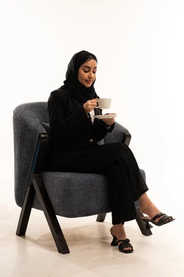 الاستمتاع بالأجواء الهادئة، احتساء المشروب المفضل, الجلوس و شرب المشروبات الساخنة والأعشاب الشتوية الدافئة, بورتيه لامرأة عربية سعودية خليجية محجبة ترتدي البدلة الرسمية السوداء تجلس على كرسي مريح و تحمل بيدها فنجان من الشاي، خلفية بيضاء