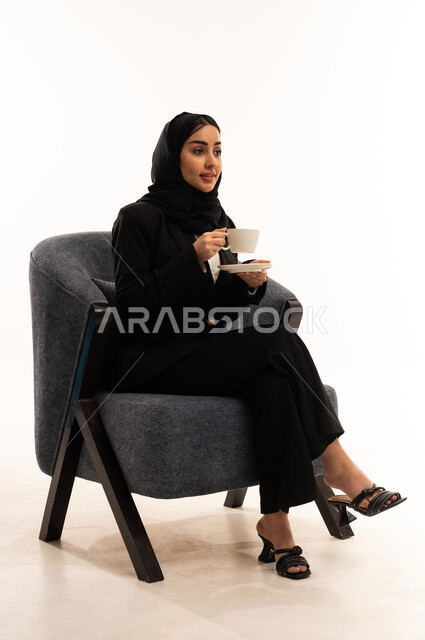 Gestures of comfort, sipping a favorite beverage, sitting and drinking hot drinks and warm winter herbs; a portrait of a veiled Saudi Arabian woman from the Gulf wearing a black formal suit, sitting in a comfortable chair and holding a cup of tea, white background.