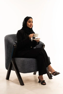 ايماءات تدل على الراحة,  احتساء المشروب المفضل, الجلوس و شرب المشروبات الساخنة والأعشاب الشتوية الدافئة, بورتيه لامرأة عربية سعودية خليجية محجبة ترتدي البدلة الرسمية السوداء تجلس على كرسي مريح و تحمل بيدها فنجان من الشاي، خلفية بيضاء