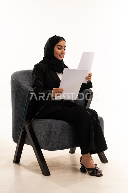 مراجعة نتائج الاجتماع، التدقيق في قراءة الملاحظات، اعداد التقارير الخاصة بالعمل، بورتيه لامرأة عربية سعودية خليجية محجبة مبتسمة ترتدي البدلة الرسمية السوداء تجلس على كرسي مريح و تحمل بيدها مجموعة من الاوراق، خلفية بيضاء