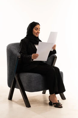 مراجعة نتائج الاجتماع، التدقيق في قراءة الملاحظات، اعداد التقارير الخاصة بالعمل، بورتيه لامرأة عربية سعودية خليجية محجبة مبتسمة ترتدي البدلة الرسمية السوداء تجلس على كرسي مريح و تحمل بيدها مجموعة من الاوراق، خلفية بيضاء