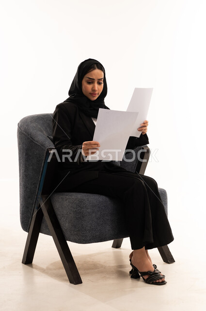 Reviewing notes, preparing work reports, a portrait of a veiled Saudi Arabian woman from the Gulf region wearing a black formal suit, sitting in a comfortable chair holding a stack of papers, reviewing and following up on plans and projects, white background.