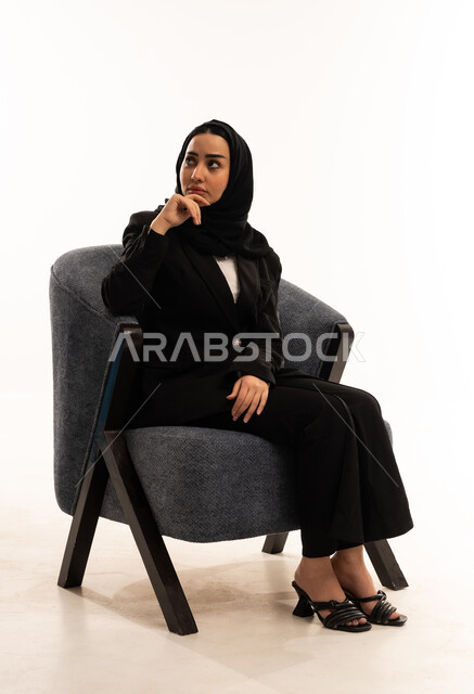 الحيرة وعدم التأكد, محاولة إيجاد حل للمشكلات الصعبة، بورتريه لامرأة عربية سعودية خليجية محجبة ترتدي البدلة الرسمية السوداء تجلس على كرسي مريح و تضع يدها على ذقنها بايماءات التفكير, مفهوم الأناقة والاعتناء بالمظهر الخارجي، خلفية بيضاء