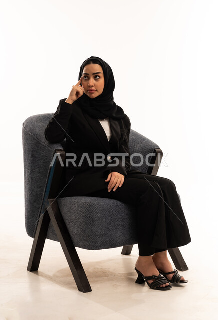 تعابير التفكير لاتخاذ القرارات, مواجهة المشكلات والتحديات في الحياة العملية، التخطيط للمستقبل و التأمل، بورتريه لامرأة عربية سعودية خليجية محجبة ترتدي البدلة الرسمية السوداء تجلس على كرسي مريح و تضع يدها على رأسها بايماءات التفكير, الاعتناء بالمظهر الخارجي، خلفية بيضاء