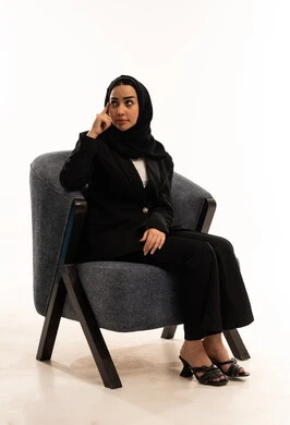 تعابير التفكير لاتخاذ القرارات, مواجهة المشكلات والتحديات في الحياة العملية، التخطيط للمستقبل و التأمل، بورتريه لامرأة عربية سعودية خليجية محجبة ترتدي البدلة الرسمية السوداء تجلس على كرسي مريح و تضع يدها على رأسها بايماءات التفكير, الاعتناء بالمظهر الخارجي، خلفية بيضاء