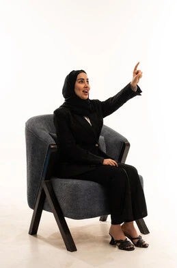 تعابير التأكد و تحديد مكان معين، الاهتمام بالمظهر الخارجي، الاشارة إلى شيء ما، ايماءات السعادة والسرور،  بورتريه لشابة عربية خليجية سعودية محجبة مبتسمة تجلس على كرسي مريح ترتدي البدلة الرسمية السوداء و ترفع اصبع السبابة للاختيار، خلفية بيضاء