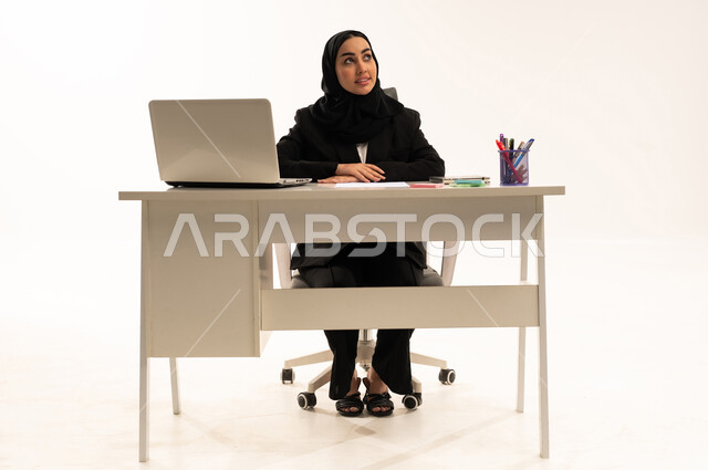 بيئة عمل مكتبيه,  تطور التكنولوجيا وإستخدام الأجهزة التقنية الحديثة, بورتريه لامرأة عربية سعودية خليجية محجبة مبتسمة تجلس خلف المكتب ترتدي البدلة الرسمية السوداء و تستخدم اللابتوب, خلفية بيضاء