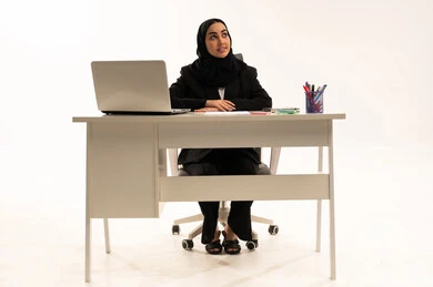 بيئة عمل مكتبيه,  تطور التكنولوجيا وإستخدام الأجهزة التقنية الحديثة, بورتريه لامرأة عربية سعودية خليجية محجبة مبتسمة تجلس خلف المكتب ترتدي البدلة الرسمية السوداء و تستخدم اللابتوب, خلفية بيضاء
