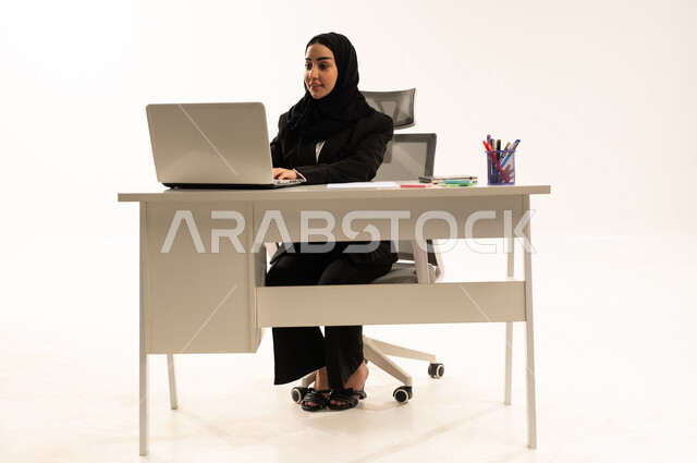 بحث وايجاد فكرة ما عبر الانترنت، ادخال البيانات من خلال لوحة المفاتيح, تطور التكنولوجيا وإستخدام الأجهزة التقنية الحديثة, بورتريه لامرأة عربية سعودية خليجية محجبة مبتسمة تجلس خلف المكتب ترتدي البدلة الرسمية السوداء و تستخدم اللابتوب, خلفية بيضاء