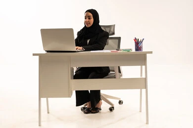 Researching and finding an idea online, data entry via keyboard, technological advancements and the use of modern devices, a portrait of a smiling, veiled Saudi Arabian woman from the Gulf region sitting behind a desk wearing a black formal suit and using a laptop, white background