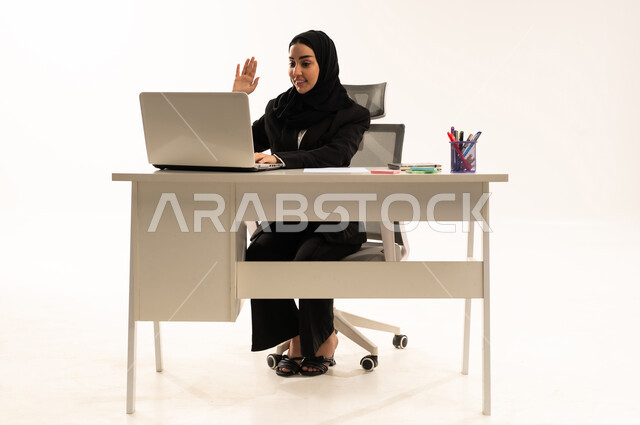 إجراء اجتماعات العمل أونلاين عبر مكالمات الفيديو، استخدام التقنيات الحديثة والتكنولوجية بالمجال الإداري، بورتريه لامرأة عربية سعودية خليجية محجبة مبتسمة تجلس خلف المكتب ترتدي البدلة الرسمية السوداء و تستخدم اللابتوب و ترفع يدها بايماءات الترحيب, خلفية بيضاء