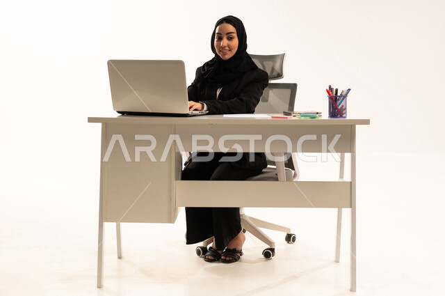 ادخال البيانات من خلال لوحة المفاتيح, تطور التكنولوجيا وإستخدام الأجهزة التقنية الحديثة, بورتريه لامرأة عربية سعودية خليجية محجبة مبتسمة تجلس خلف المكتب ترتدي البدلة الرسمية السوداء و تستخدم اللابتوب, خلفية بيضاء
