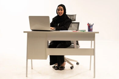 Data entry via keyboard, technological advancements and the use of modern devices, portrait of a smiling, veiled Saudi Arabian woman from the Gulf region sitting behind a desk wearing a black formal suit and using a laptop, white background