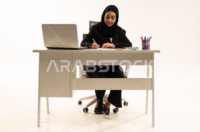ملفات ووثائق خاصة بالعمل, إجراء الاعمال المكتبيه داخل مقر الشركه، بورتريه لامرأة عربية سعودية خليجية محجبة ترتدي البدلة الرسمية السوداء تجلس خلف المكتب و أمامها جهاز اللابتوب، دراسه المشاريع ومراجعه التقارير السنويه، خلفية بيضاء
