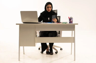 Busy at work, sending emails via computer, communicating with clients remotely, a portrait of a veiled Saudi Arabian woman wearing a black formal suit, using a laptop and holding a mobile phone, white background